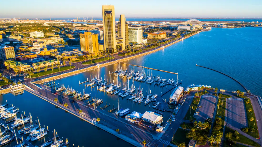 Sunlit aerial view of a marina with boats, a tall building, and a bridge over the water - things to do south texas