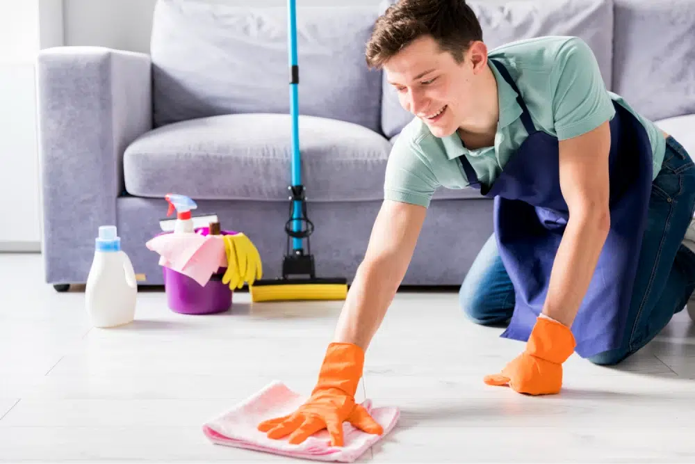 Man cleaning the floor with a pink cloth while smiling, cleaning supplies in the background.