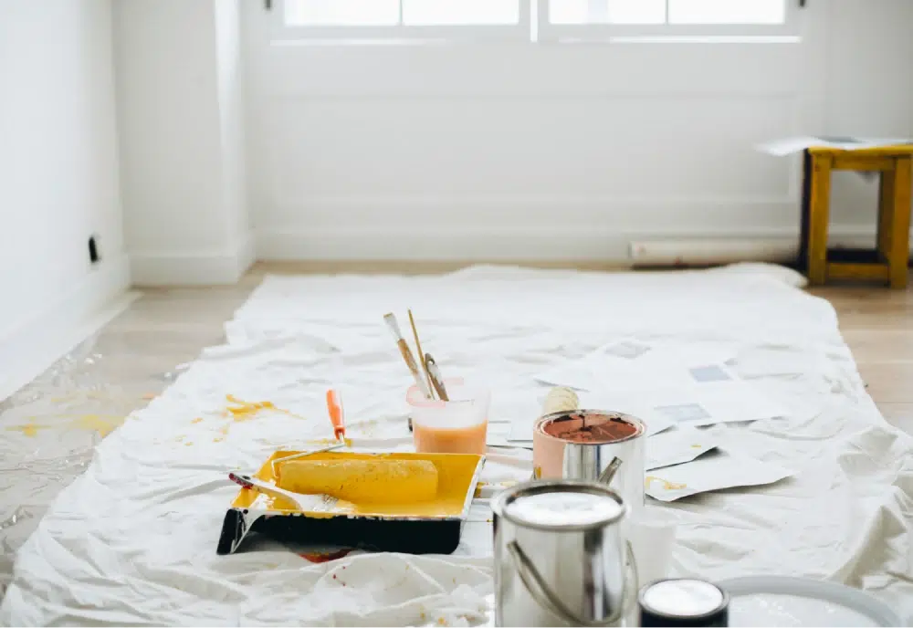 Painting supplies including a roller, brushes, and paint cans on a white sheet in a room under renovation.